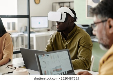 Man immersed in virtual reality using VR headset while sitting in contemporary office space alongside two colleagues collaborating on digital devices