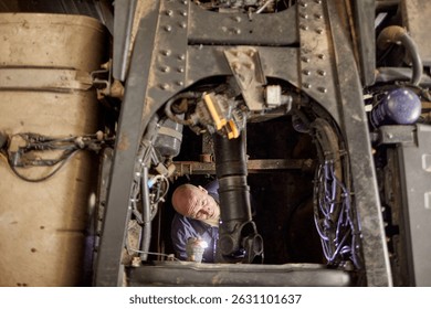 Man inspecting complex machinery in industrial setting, holding flashlight for better visibility. Detailing mechanical parts and working environment, emphasizing maintenance process