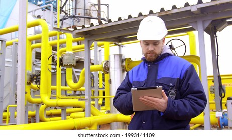 Man Inspector Using Digital Tablet At Gas Station. Portrait Of Engineer In Safety Hardhat And Uniform Supervising Large Industrial Gas Pipeline