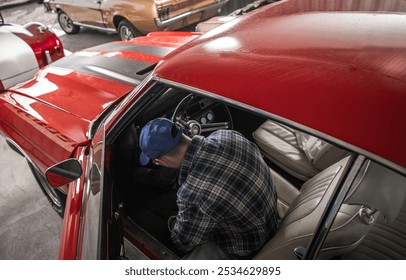 A man inspects the interior of a classic red car, focusing intently on the dashboard while surrounded by other vintage automobiles in a well-lit garage. - Powered by Shutterstock