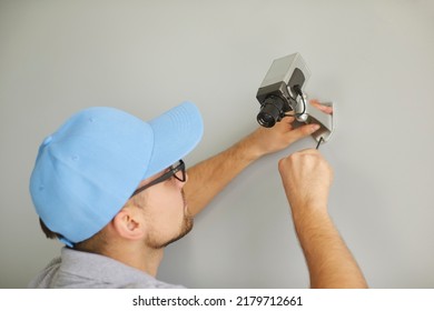 Man Installing CCTV Surveillance Camera Inside Building. Young Male Worker In Blue Mockup Mock Up Company Uniform Baseball Cap Hat Uses Screwdriver While Setting Up New Modern Security Camera On Wall