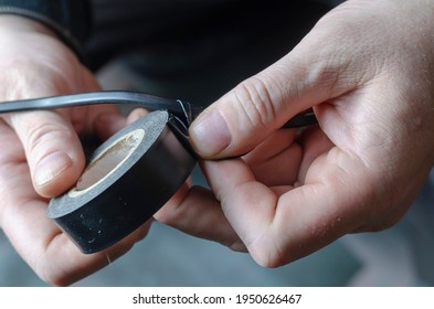 A Man Is Insulating A Black Electrical Wire. Close-up Of Male Hands Holding A Roll Of Black Duct Tape For Electrical Work. Adult Male, Caucasian. Inside The Room. Selective Focus.