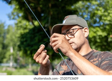 A man intently tying a fishing line to the rod in an outdoor park setting. He wears a camouflage shirt and cap, demonstrating concentration and skill in his fishing preparation. - Powered by Shutterstock