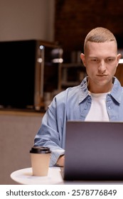 Man intently using laptop while sitting in cafe with coffee cup nearby, appearing deeply focused on work or study, wearing casual denim shirt and white T-shirt