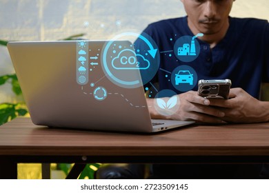 A man interacts with a smartphone while working on a laptop, surrounded by digital graphics representing environmental impact. The scene highlights technology's role in sustainability awareness.