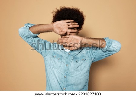 Man introvert hiding face behind opened palms, feeling scared or ashamed of his mistakes and behavior, dressed in denim clothes isolated on brown studio background. Embarrassment and shyness