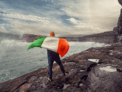Man With Irish Flag Standing On The Edge Of A Cliff, Stunning Scenery With Ocean, Cliff And Cloudy Sky And Fog Over Water In The Background. Aran Island, County Galway, Ireland. Travel Theme.