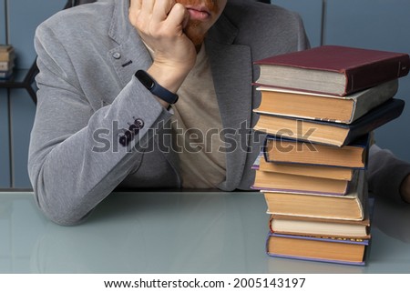 the man in the jacket has his head propped up and he's looking at a large stack of books. hand and books in the frame