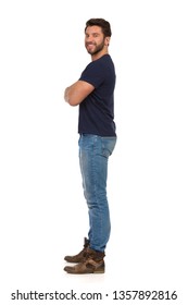 Man In Jeans And Blue T-shirt Is Standing With Arms Crossed, Looking At Camera And Smiling. Side View. Full Length Studio Shot Isolated On White.