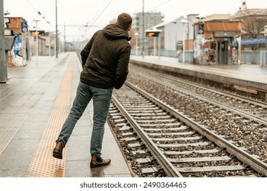 A man in jeans and a brown jacket stands on a train platform, looking down the tracks. - Powered by Shutterstock