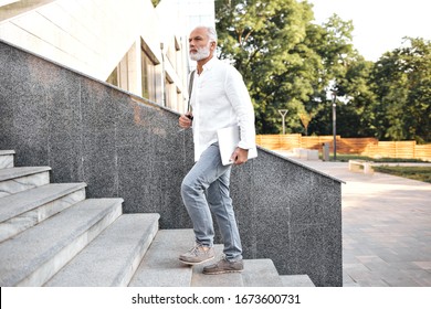 Man In Jeans And Shirt Goes Upstairs And Holds Laptop. Attractive Adult With Gray Beard In Office Clothes Is Walking Up Gray Stairs Outside..