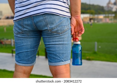 man in jeans shorts and striped T-shirt with blue bottle of water in right hand on field background