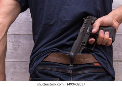 A Man In Jeans And A T-shirt Holds A Gun In His Hand From The Back