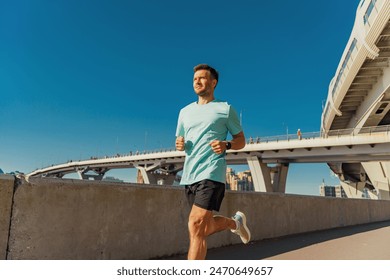 A man jogs along a path under a clear blue sky, with a bridge and urban buildings in the background.