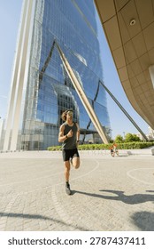 A man jogs on a paved path next to a tall, glass building during the day. The sky is clear, and the area is busy with greenery and visible structures.