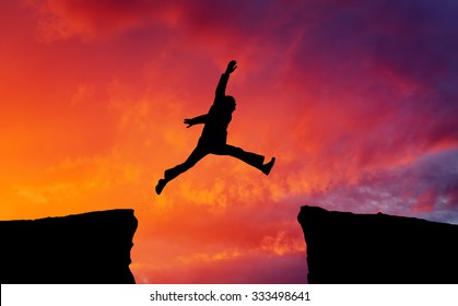 Man jumping across the gap from one rock to cling to the other. Man jumping over rocks with gap on sunset fiery background. Element of design.  