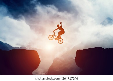 Man jumping on bmx bike over precipice in mountains at sunset. Raising hand showing hello gesture. Extreme sport, risk, cycling. - Powered by Shutterstock