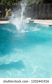 Man Jumps Into An Outdoor Swimming Pool In The Summer. Man Lands In Pool From Jumping Off Diving Board Doing Cannon Ball Into A Pool With Big Splash As He Lands In The Pool.