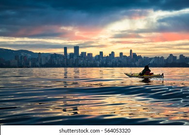 Man Kayaking In Front Of Downtown Vancouver, BC, Canada. Picture Taken During A Cloudy Winter Sunrise.