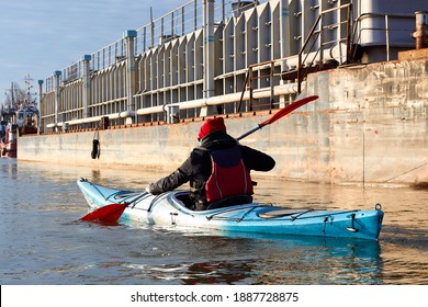 Man Kayaking On Blue Kayak In Winter Danube River Near An Old Rusty Barge. Back View.