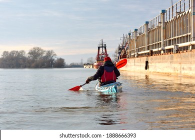 Man Kayaking On Blue Kayak In Winter Danube River Near An Old Rusty Barge. Back View.