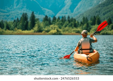 A man kayaking on a calm lake, with a backdrop of lush green trees and a distant mountain range. The water is clear and blue, reflecting the sunny sky. He is wearing a life jacket - Powered by Shutterstock