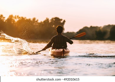 Man kayaking. Rear view of young man splashing water while kayaking on river with sunset in the background - Powered by Shutterstock