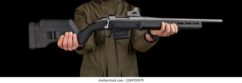 A Man In A Khaki Jacket Holds A Bolt Rifle With A Reflex Sight In His Hands. Hunter With Weapons. Dark Background.