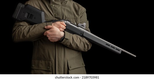 A Man In A Khaki Jacket Holds A Bolt Rifle With A Reflex Sight In His Hands. Hunter With Weapons. Dark Background.