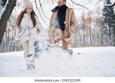 Man is kicking the snow. Happy couple having a walk in winter forest. Blue car is parked. - Powered by Shutterstock