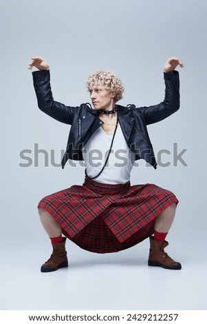 Man in kilt and leather jacket posing with raised arms, embodying a mix of traditional and punk style against grey studio background. Concept of fashion fusion, art, uniqueness, self-expression. Ad