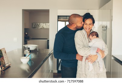 Man Kissing His Wife Holding A Newborn Baby Boy In Kitchen. Lovely Young Family Of Three In Morning In Kitchen.