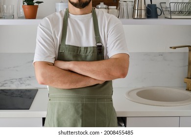 A Man In A Kitchen Apron Stands In A Modern Kitchen. Cooking At Home In Uniform, Protection Apparel. Green Fabric Apron, Casual Clothing. A Man In An Apron Prepares To Prepare A Meal