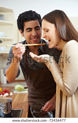 Man in kitchen letting woman taste a soup with a wooden spoon
