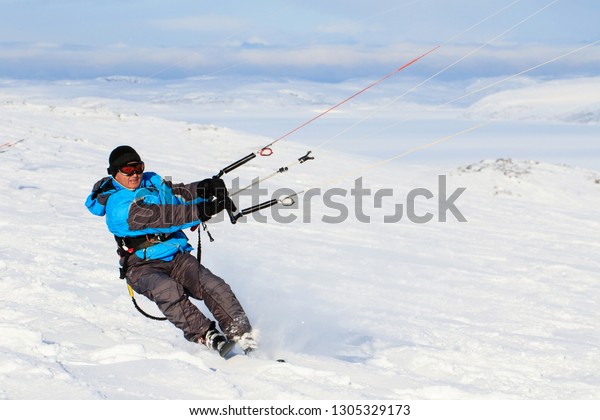 Man kite boarding rider sportsman with kite in\
sky rides in snow on kite board. Recreational activity, extreme\
active sports, snow kiting ski, outdoor, winter snowy season,\
skiing. Blue sky background