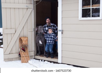 Man Kneeling Next To Happy Smiling Young Son And Very Tall And Muscular Male Blue Great Dane In The Open Door Of A Garden Shed In Winter