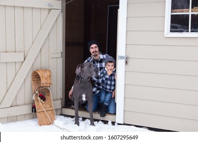 Man Kneeling Next To Young Son And Very Tall And Muscular Male Blue Great Dane In The Open Door Of A Garden Shed In Winter