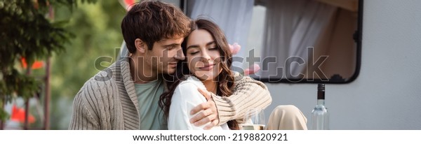 Man in knitted cardigan\
hugging girlfriend near glass of wine and camper van outdoors,\
banner