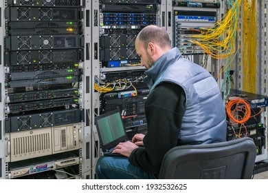 A man with a laptop sits in a server room. A technician works near the racks of a modern data center. The system administrator configures the computer hardware.