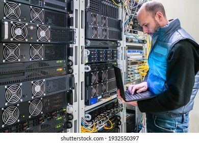 A Man With A Laptop Stands In The Server Room. A Technician Works Near The Racks Of A Modern Data Center. The System Administrator Configures The Computer Hardware.