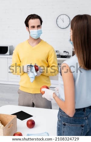 Man in latex gloves cleaning apple near digital tablet and blurred girlfriend in medical mask in kitchen