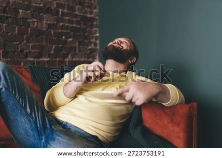 Man laughs freely while leaning back on couch, holding a chip, expressing pure amusement and relaxed energy during solo snack time, unplugged from screens and tech devices