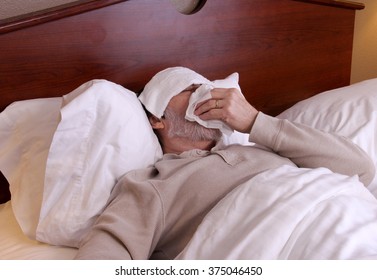 Man Laying In Bed With A Washcloth On His Forehead And Blowing His Nose