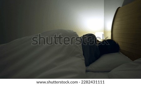 Man laying down in bed to rest after long day. Person going to sleep going under bedsheets resting turning bedside lamp off