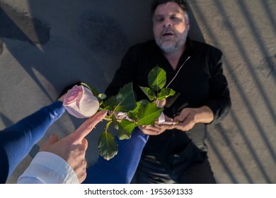 Man Laying On The Ground With A Pink Rose In His Hands, While A Woman Stands Above Him And Points A Finger At Him, POV Shot