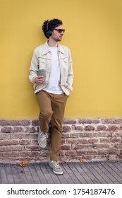 man leaning against a yellow wall on the street drinking coffee and listening to music