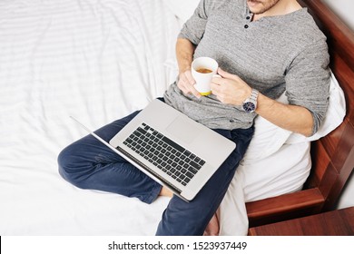 Man Leaning On Big Comfy Chair On His Bed, Drinking Cup Of Coffee And Watching Movie Or Webinar On Laptop