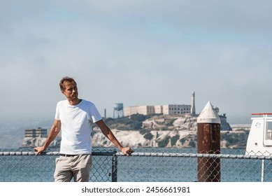 Man leaning on metal fence with hands on railing, overlooking Alcatraz Island in San Francisco. Clear sky, relaxed tourist vibe, sightseeing day out. - Powered by Shutterstock