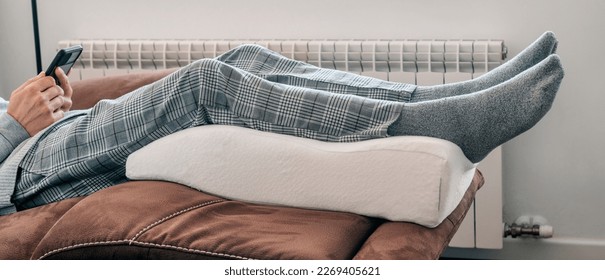 a man leans his legs on a leg elevation pillow made of memory foam, resting on the sofa, in a panoramic format to use as web banner or header