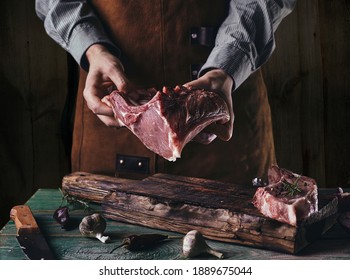 A Man In A Leather Apron Holds A Piece Of Raw Meat With A Bone. Pork Ribs In The Hands Of The Butcher.